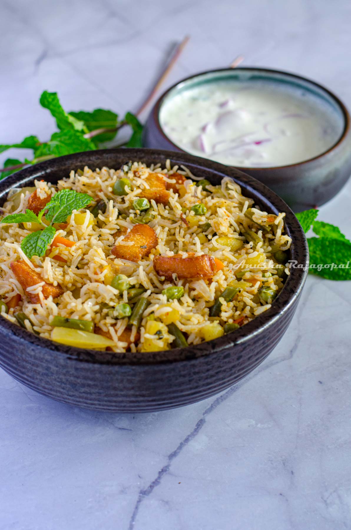 Brinji rice served in a black bow. Mint leaves by the side and chilled onion raita at the background.