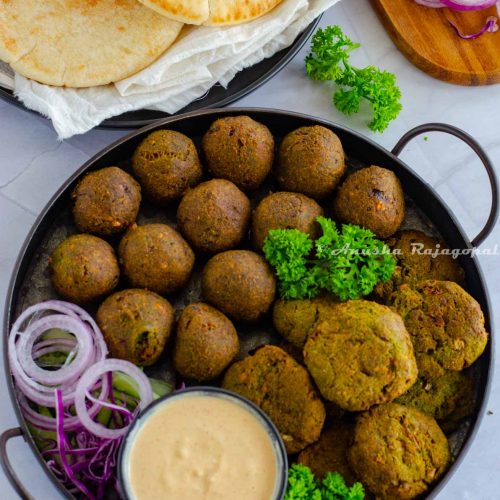 falafels arranged on a serving tray with onion rings, herbs and a dip. Pita bread and other chopped veggies at the background.