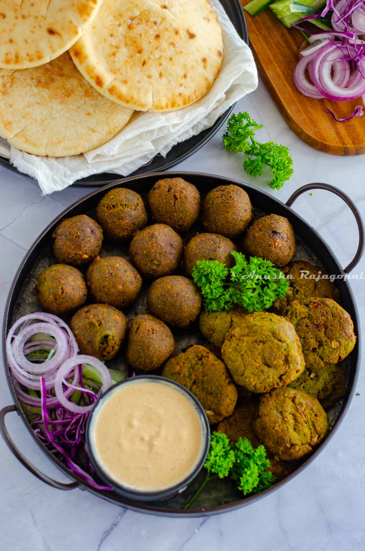 falafels arranged on a serving tray with onion rings, herbs and a dip. Pita bread and other chopped veggies at the background.