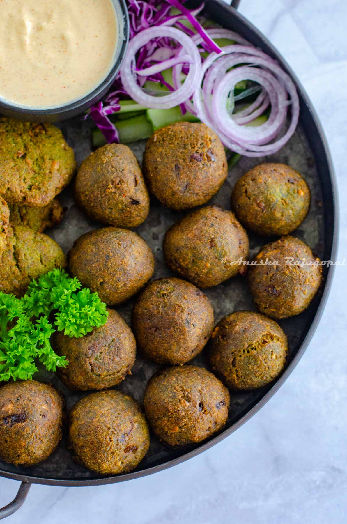 deep fried falafels arranged on a metal serving tray with parsley, a creamy dip and chopped veggies