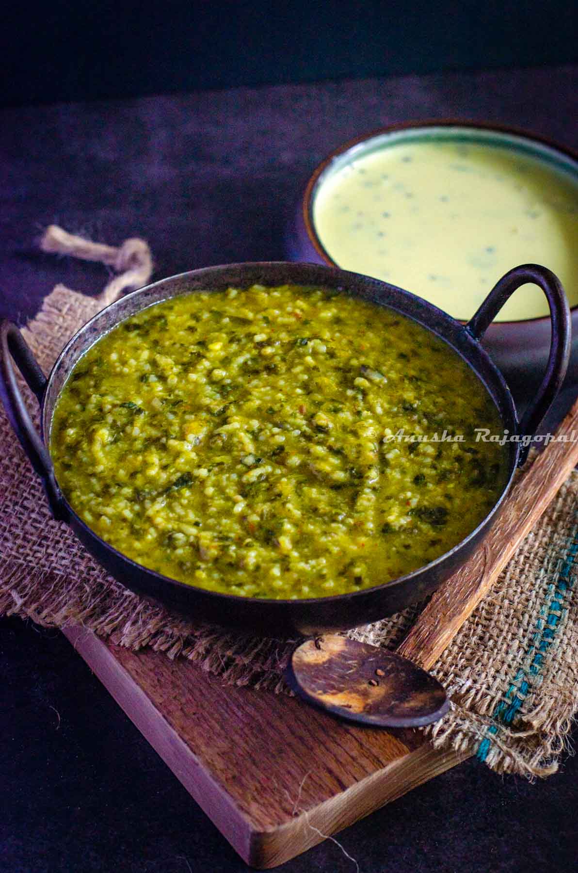 creamy palak khichdi served in an iron wok placed over a burlap mat on a wooden chopping board. Gujarati Kadhi served as an accompaniment. Wooden spoon by the side of the wok.