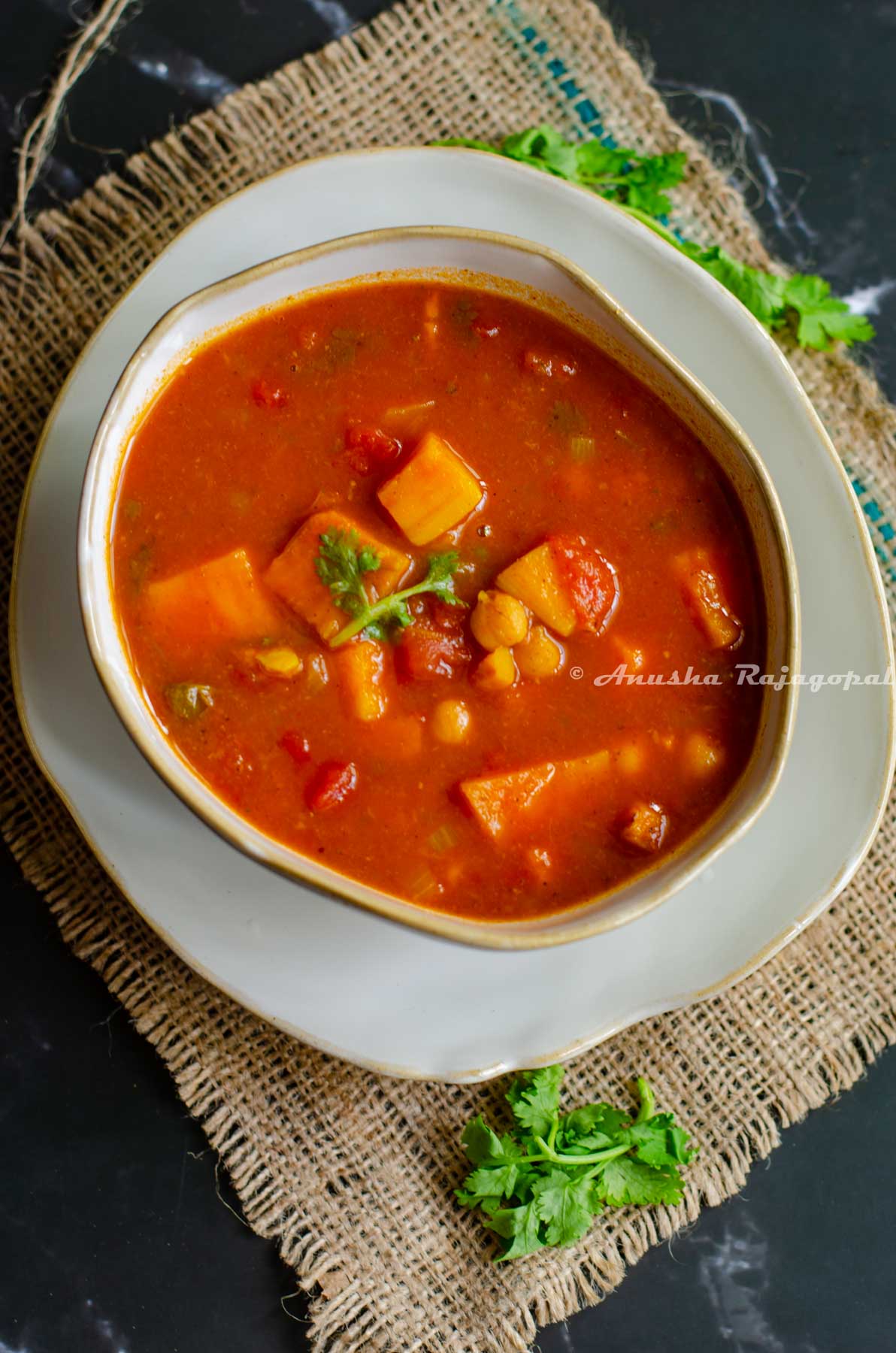 vegan sweet potato and chickpea curry serve in an asymmetrical bowl placed over a similar asymmetrical deep plate. Kept on a burlap mat against a black backrdrop. Cilantro leaves scattered by the side.