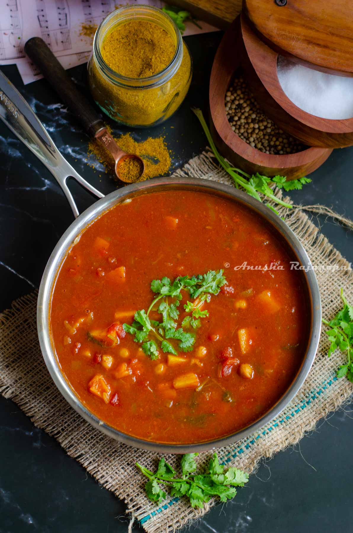 chickpeas and sweet potato curry served in a stainless steel skillet placed over a burlap mat. Curry powder in a jar, salt cellar and cilantro around the skillet