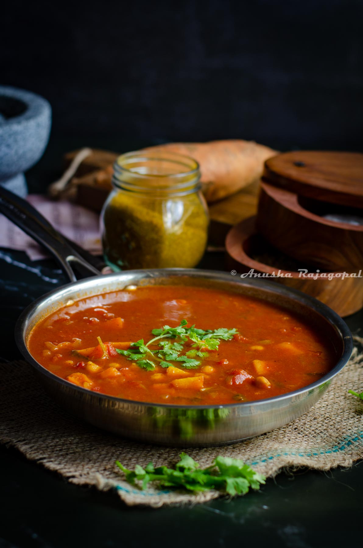 chickpeas and sweet potato curry served in a stainless steel skillet placed over a burlap mat. Curry powder in a jar, salt cellar and cilantro around the skillet