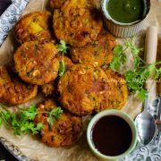 air fryer aloo paneer tikkis served over a rustic serving tray lined with unbleached parchment paper. Green chutney and sweet chutney in two small bowls placed on the platter. Cilantro leaves as garnish and a spoon by the side.