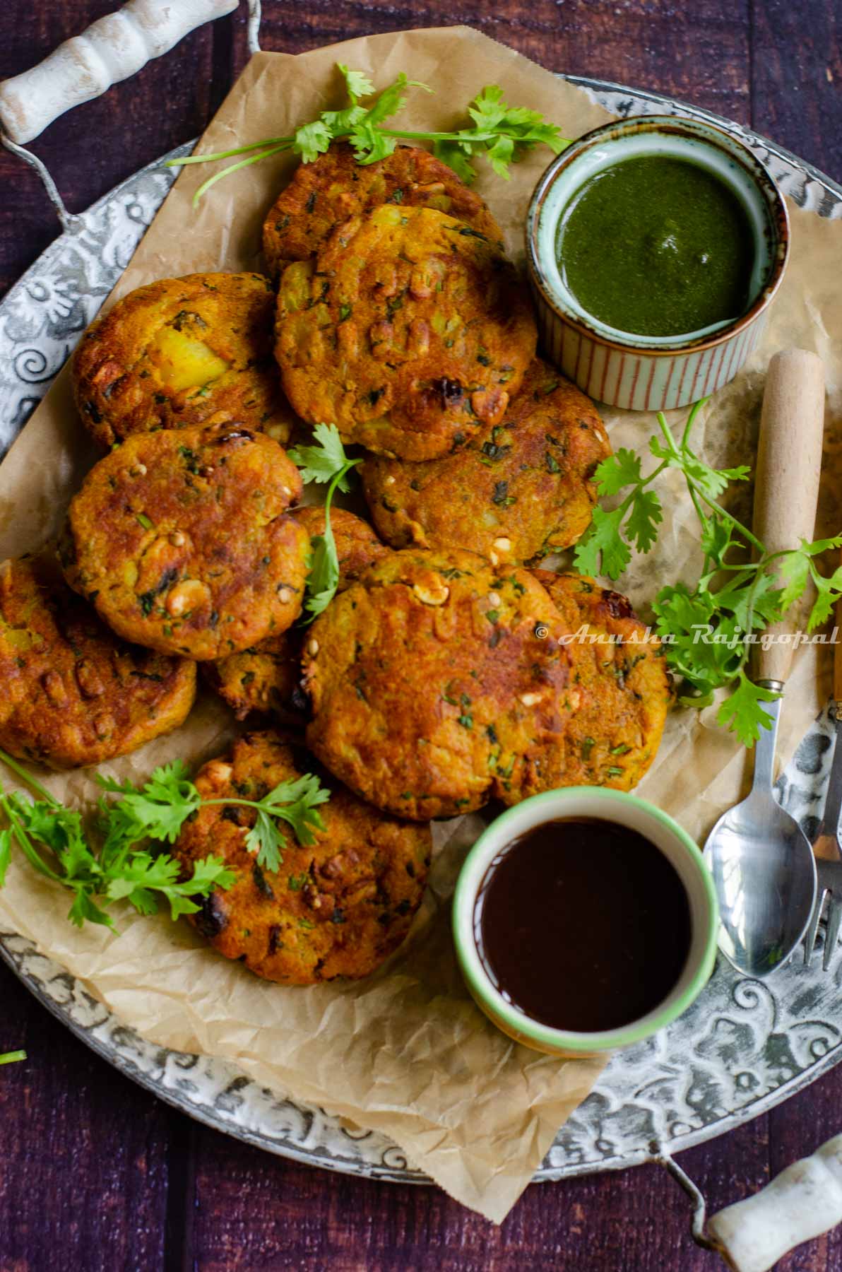 air fryer aloo paneer tikkis served over a rustic serving tray lined with unbleached parchment paper. Green chutney and sweet chutney in two small bowls placed on the platter. Cilantro leaves as garnish and a spoon by the side.