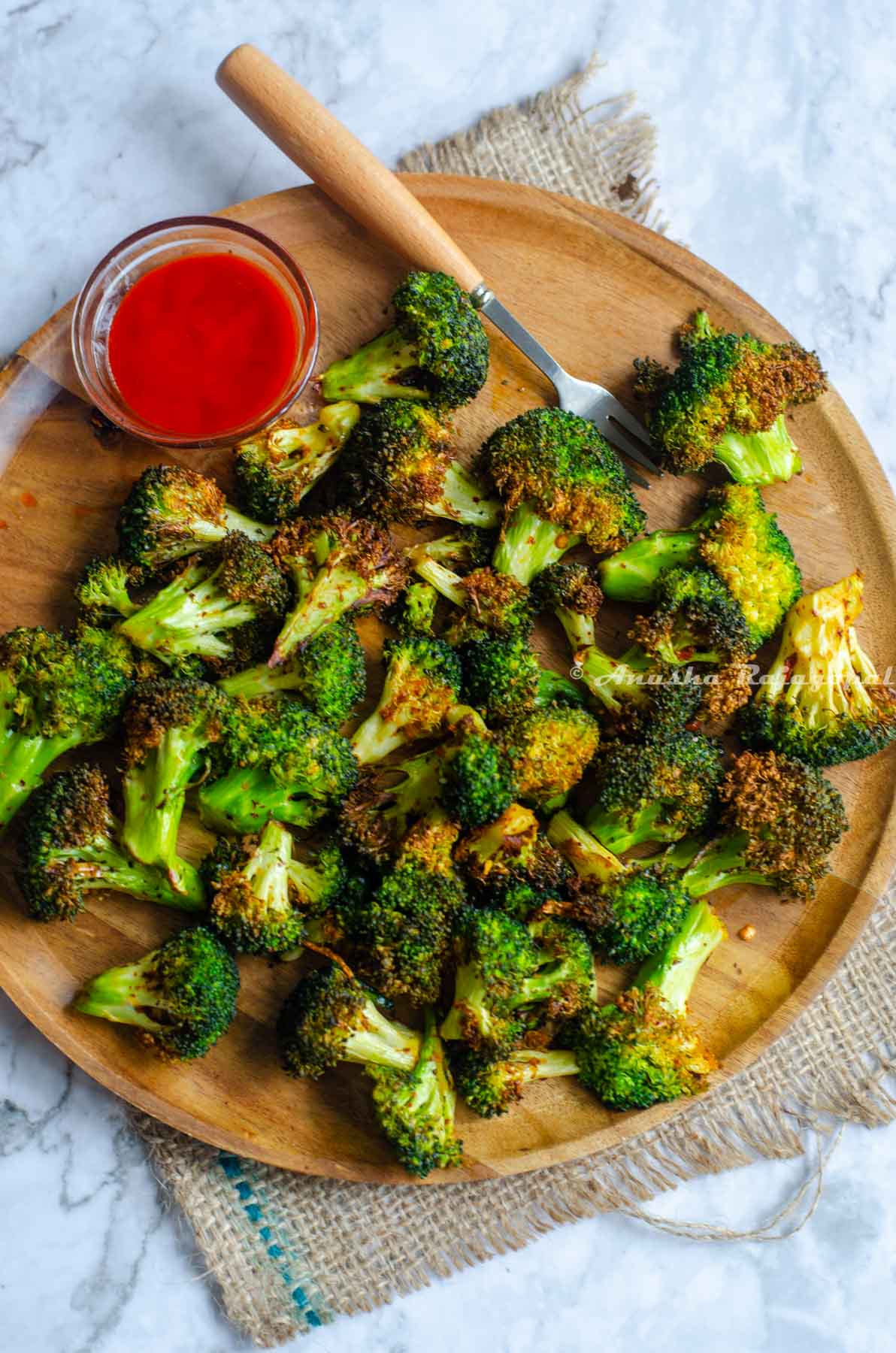 air fried broccoli served on a round wooden plate with a small pinch bowl of sriracha sauce and a wooden handled steel spoon