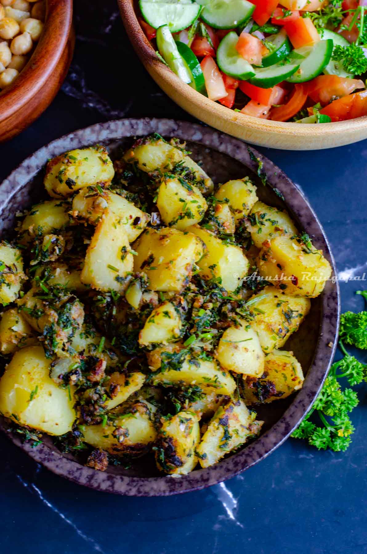 Batata Harra served in a shallow black bowl placed over a black marble background. Parsley by the side as garnish.