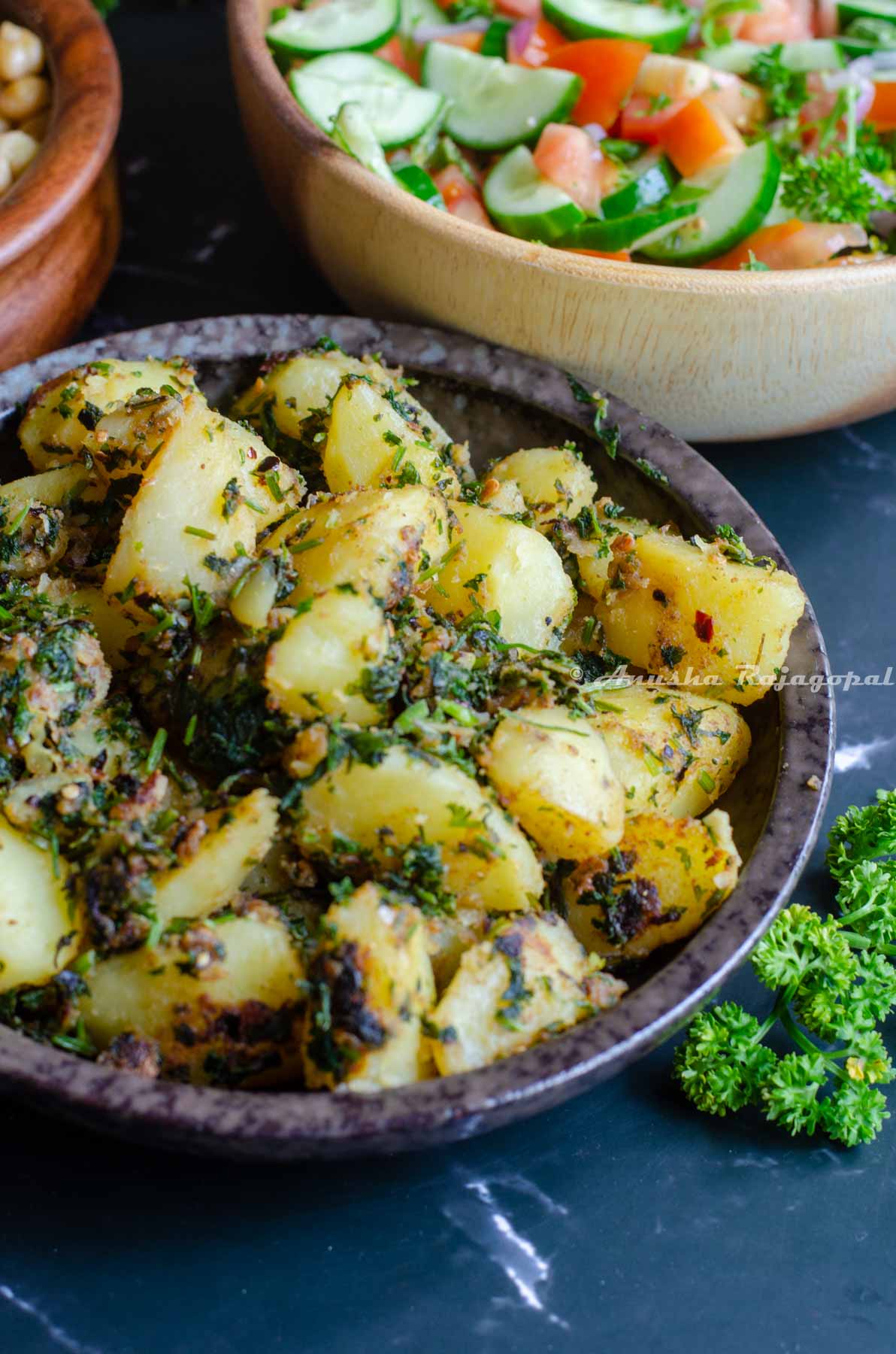 Batata Harra served in a shallow black bowl placed over a black marble background. Parsley by the side as garnish.