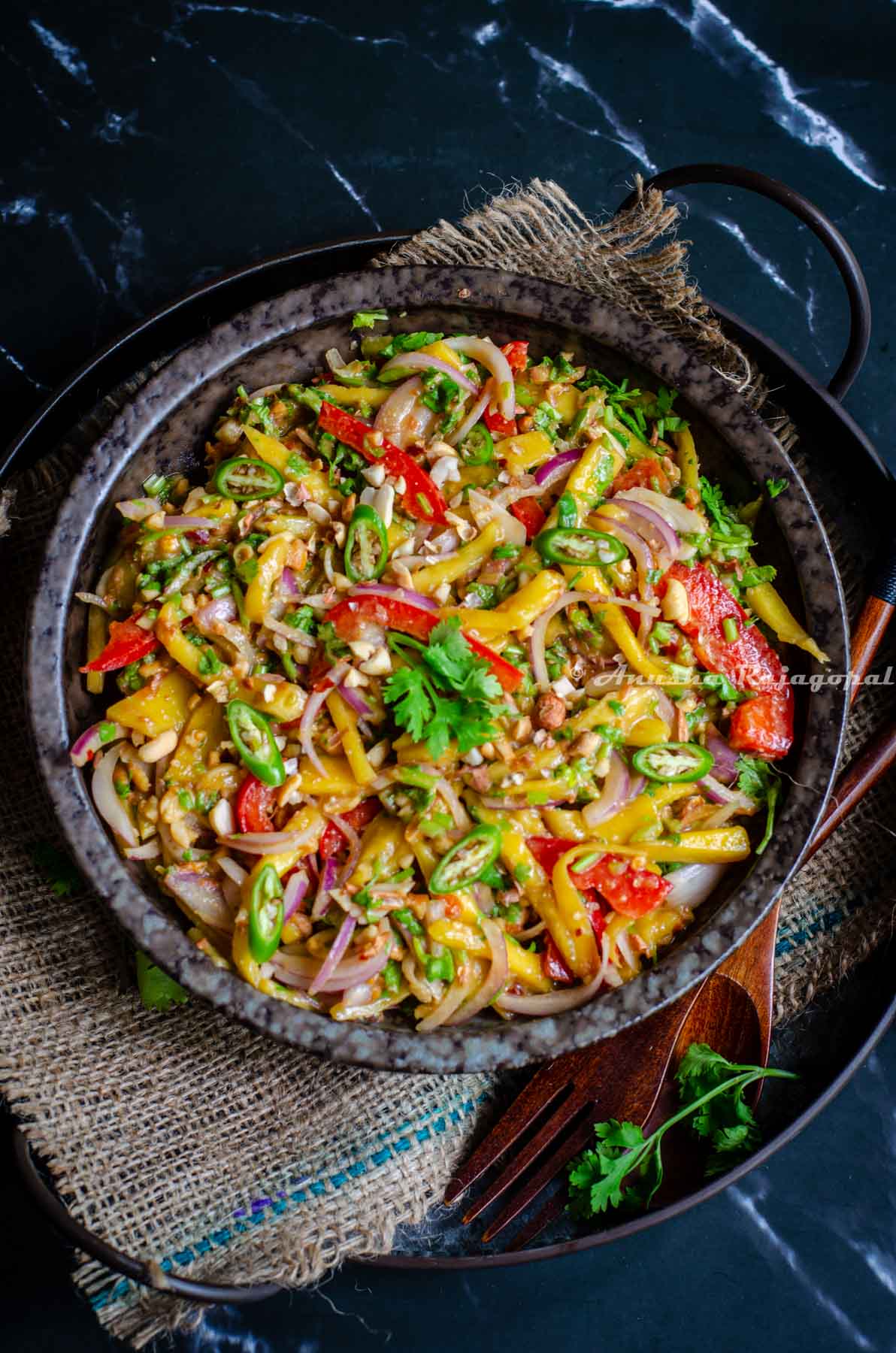 A sweet and spicy mango salad made the Thai way and served in a black shallow bowl placed over a burlap mat. Spoon and fork by the side