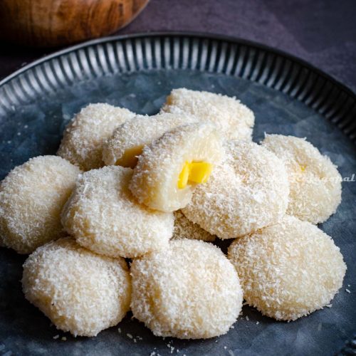 Mango stuffed mochis stacked and served on a rustic metal platter. Chopped mangoes in a small wooden bowl at the background