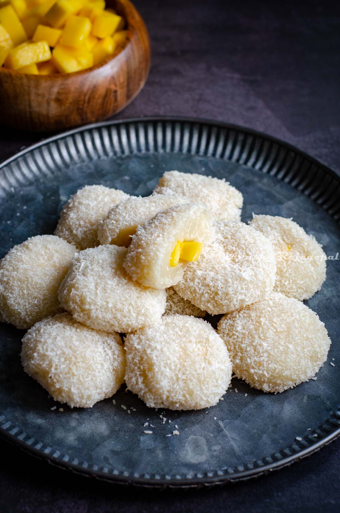 Mango stuffed mochis stacked and served on a rustic metal platter. Chopped mangoes in a small wooden bowl at the background