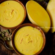 two servings of mango shrikhand served in brown ceramic bowls placed on a wooden tray. Cardamom and saffron along with cut mangoes by the side.