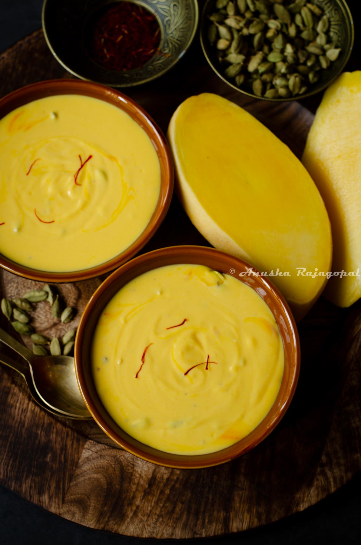 two servings of mango shrikhand served in brown ceramic bowls placed on a wooden tray. Cardamom and saffron along with cut mangoes by the side.