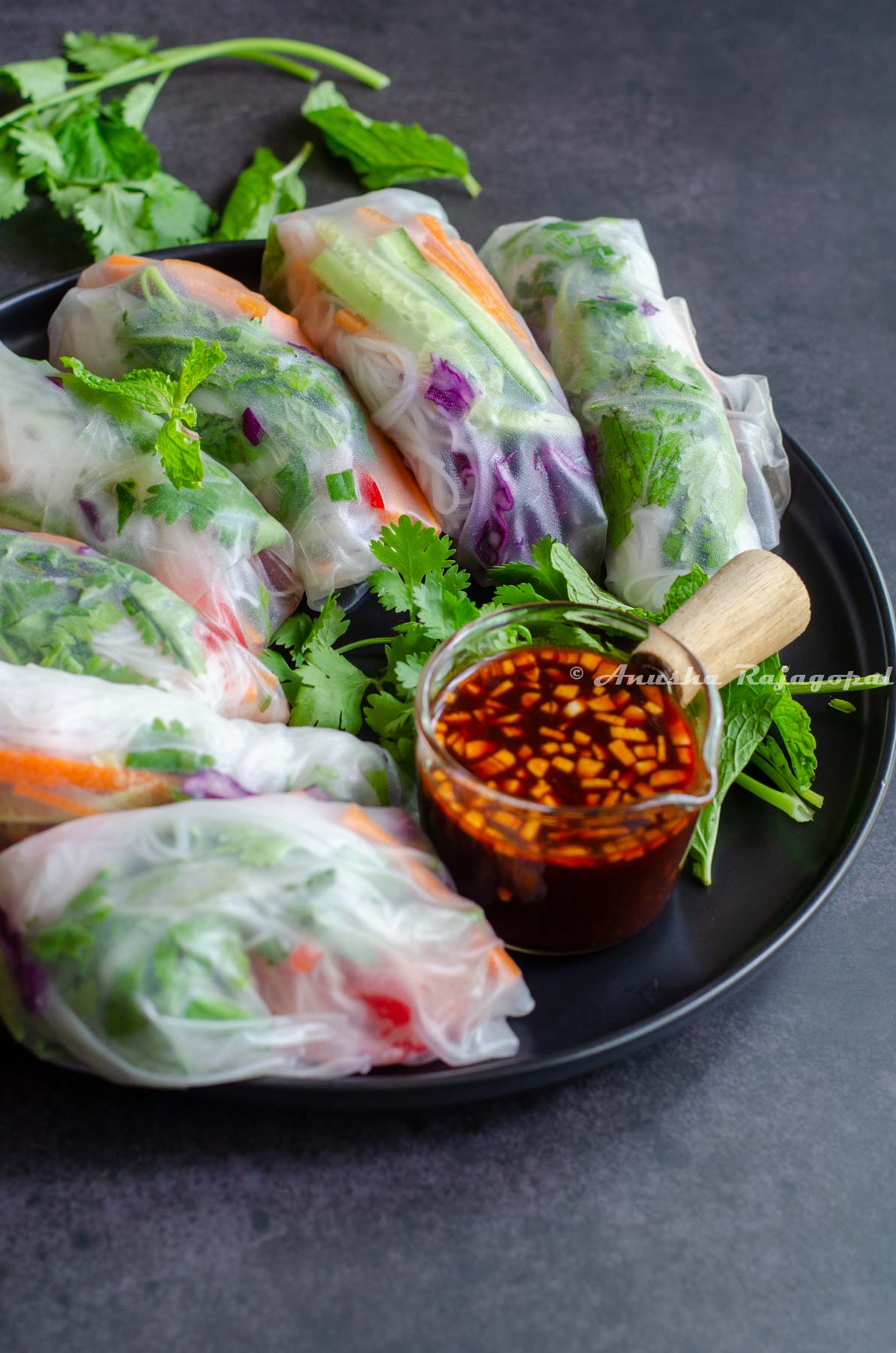 vegan summer rolls served on a black dinner plate with a small bowl of spicy dipping sauce speckled with chopped garlic.