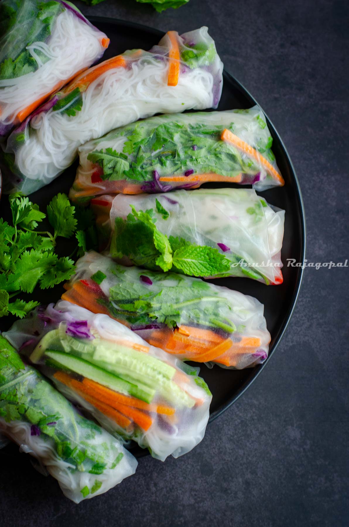 summer rolls wrapped and placed on a black plate over a black background. Fresh cilantro in the middle of the plate but appears blurred.