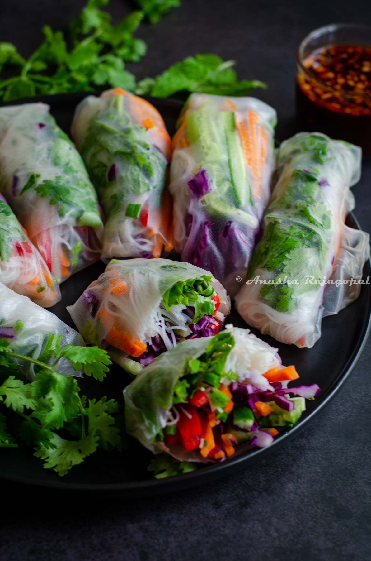 vegan summer rolls served on a black plate. One roll has been cut into halves to show the contents. A small glass bowl of dipping sauce at the back and herbs scattered at the back of the plate.