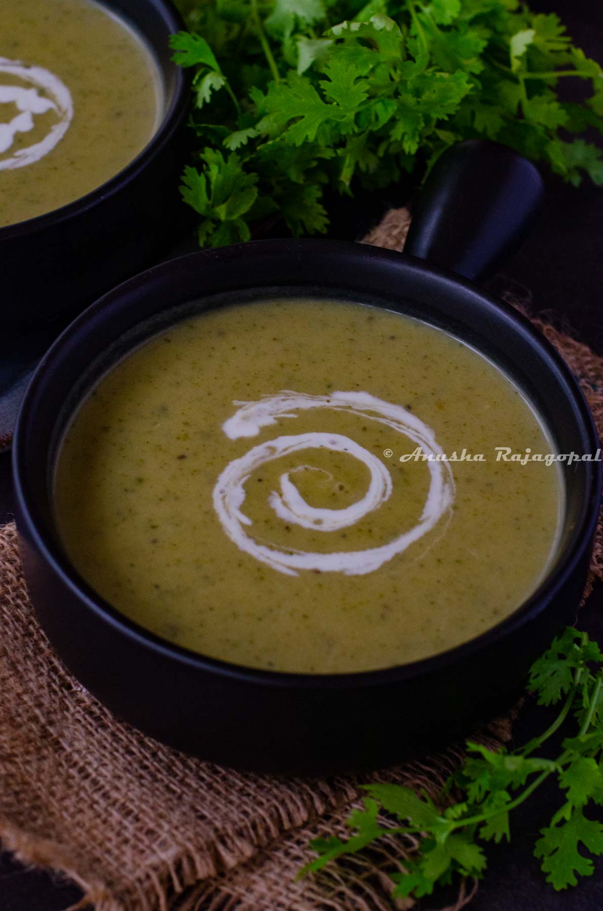 vegan zucchini soup served in a handled black soup bowl. Coconut milk swirled on the soup for aesthetics. Herbs at the background. The bowl has been placed over a burlap mat.