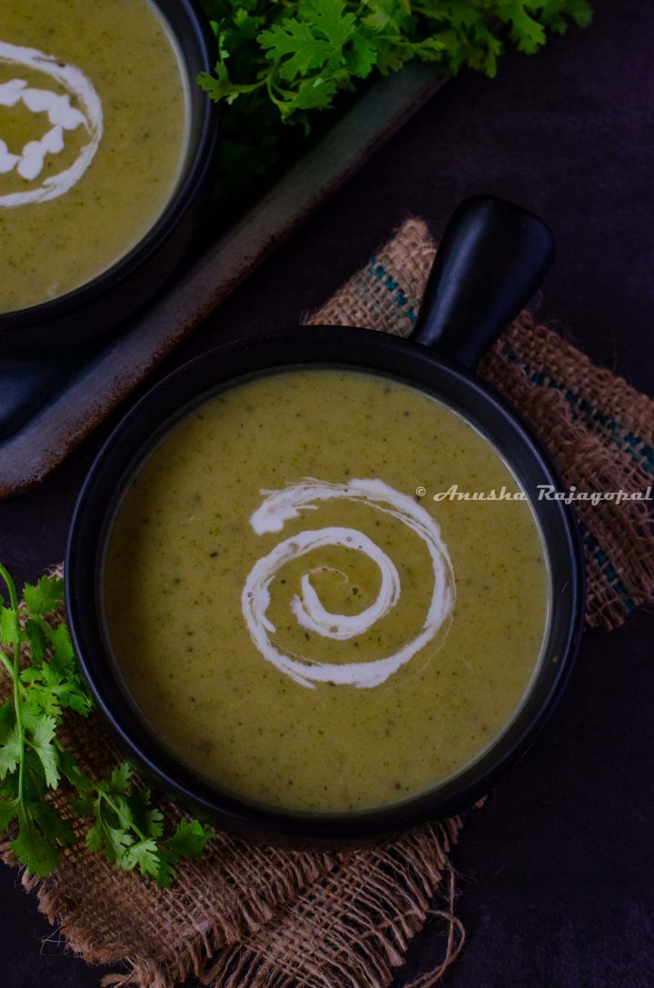 vegan zucchini soup served in a handled black soup bowl. Coconut milk swirled on the soup for aesthetics. Herbs at the background. The bowl has been placed over a burlap mat.