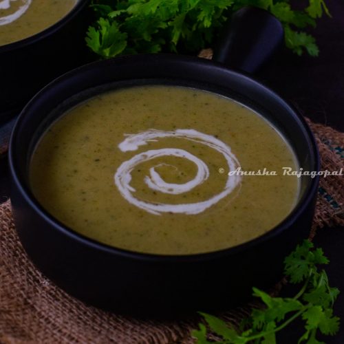 vegan zucchini soup served in a handled black soup bowl. Coconut milk swirled on the soup for aesthetics. Herbs at the background. The bowl has been placed over a burlap mat.