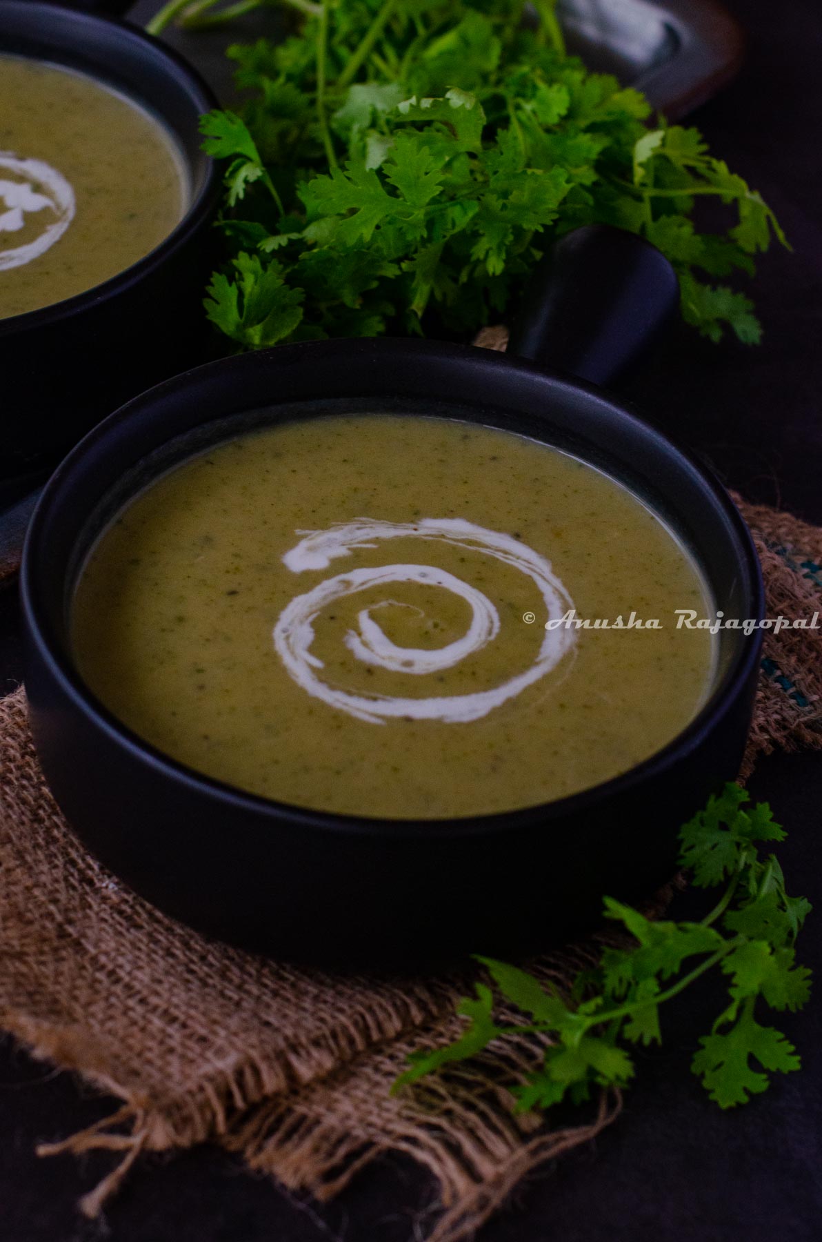 vegan zucchini soup served in a handled black soup bowl. Coconut milk swirled on the soup for aesthetics. Herbs at the background. The bowl has been placed over a burlap mat.