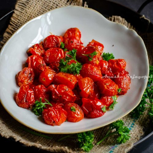 roasted cherry tomatoes served in a beige shallow plate with an uneven rim. The plate is placed on a burlap mat over an iron serving tray against a black marbled backdrop.