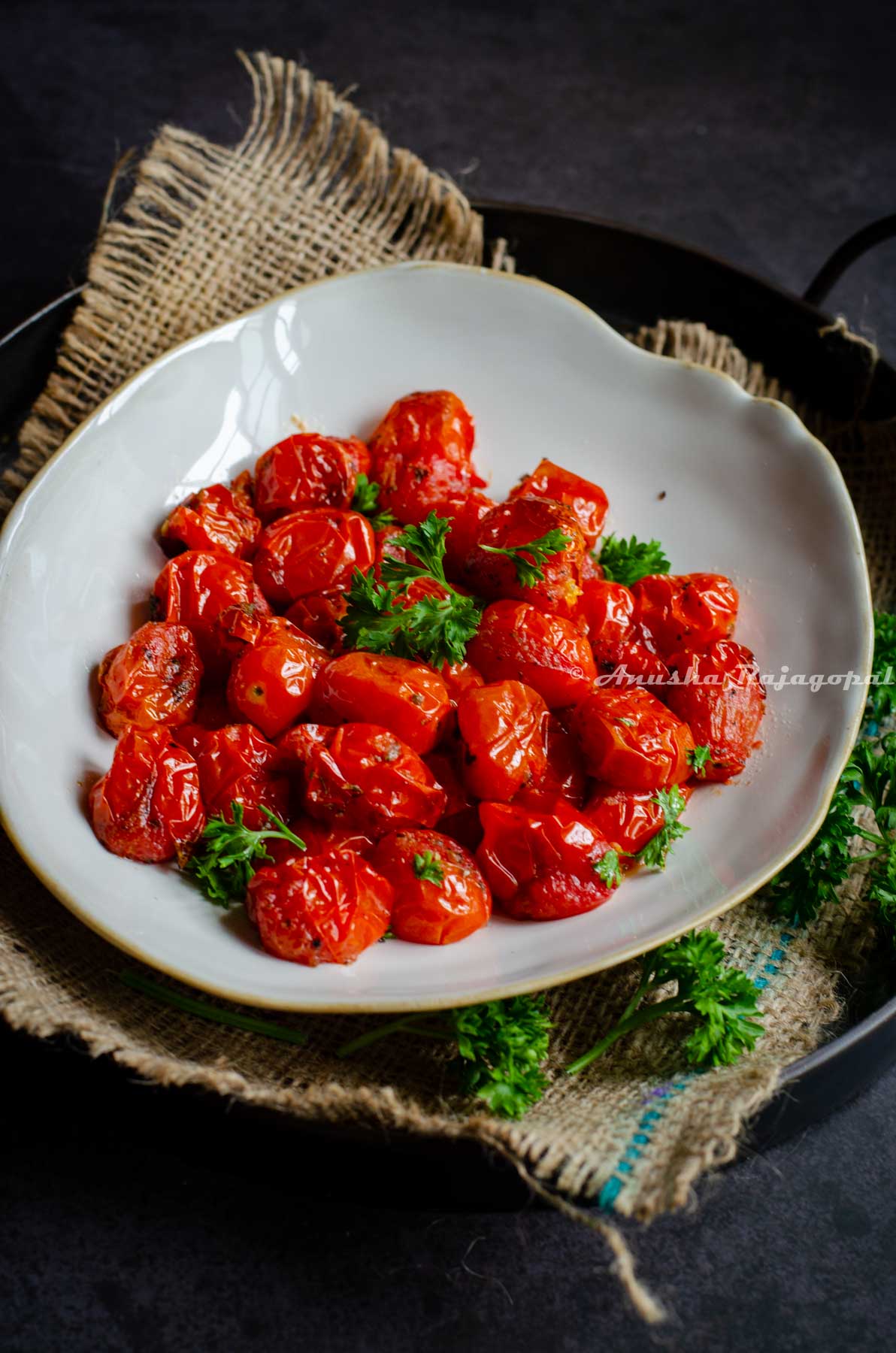 roasted cherry tomatoes served in a beige shallow plate with an uneven rim. The plate is placed on a burlap mat over an iron serving tray against a black marbled backdrop.