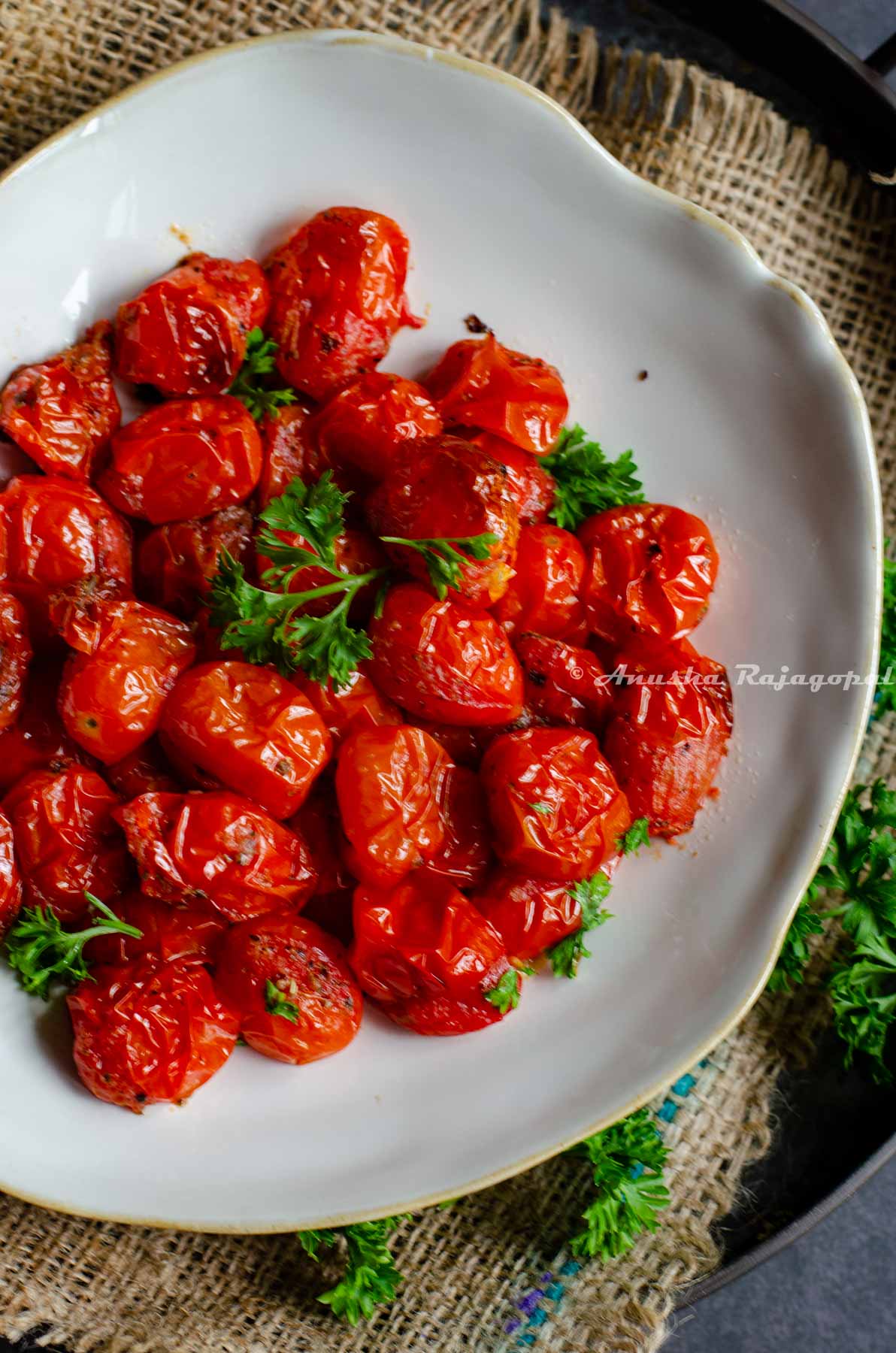 air fryer tomatoes served in a beige shallow plate with an uneven rim. The plate is placed on a burlap mat over an iron serving tray against a black marbled backdrop.