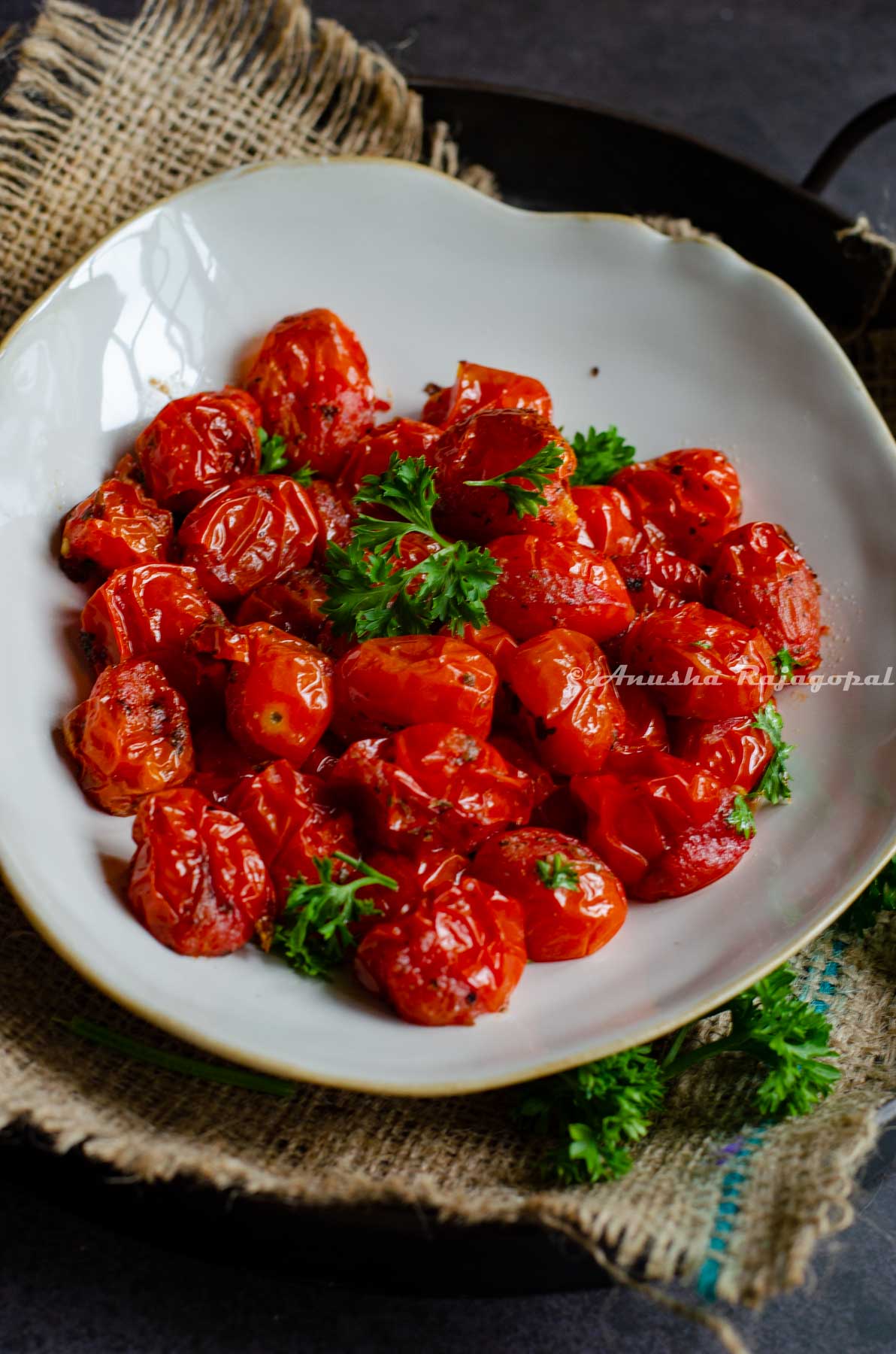 roasted cherry tomatoes served in a beige shallow plate with an uneven rim. The plate is placed on a burlap mat over an iron serving tray against a black marbled backdrop.