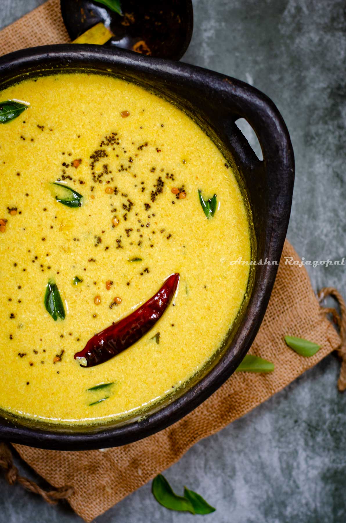Kerala style mango and yogurt gravy served in a black clay frying pan. Pan placed over a burlap mat . Curry leaves strewn around. A wooden ladle at the back.