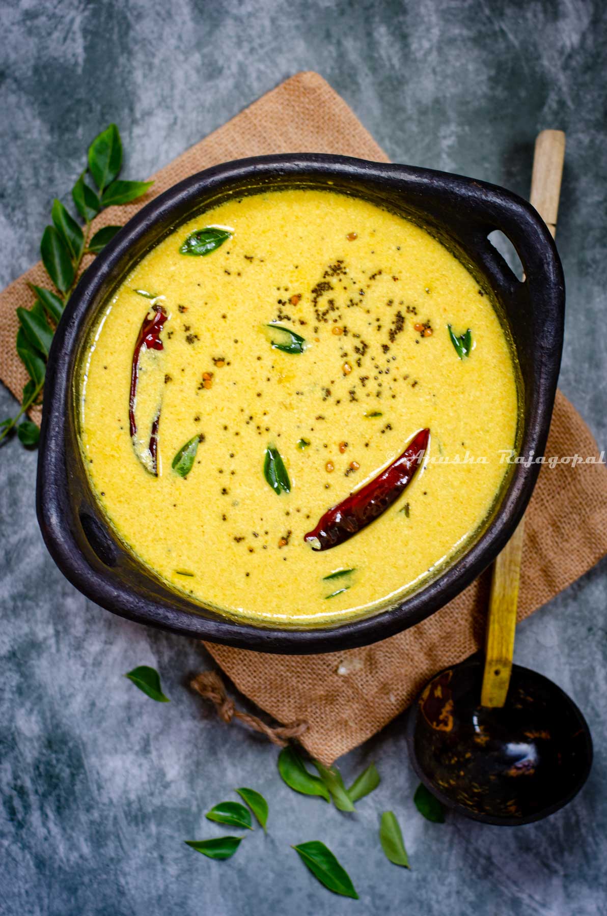 Kerala style mango and yogurt gravy served in a black clay frying pan. Pan placed over a burlap mat . Curry leaves strewn around. A wooden ladle at the back.