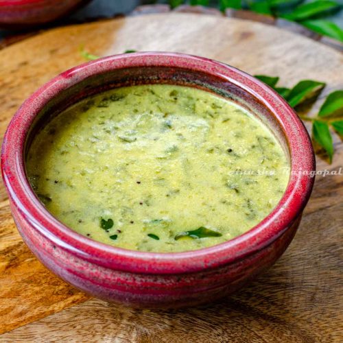 South Indian Mor Kulambu served in a reddish bowl placed over a wooden platter. Curry leaves at the background.