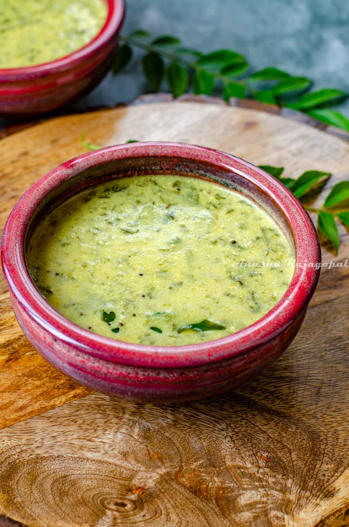 South Indian Mor Kulambu served in a reddish bowl placed over a wooden platter. Curry leaves at the background.