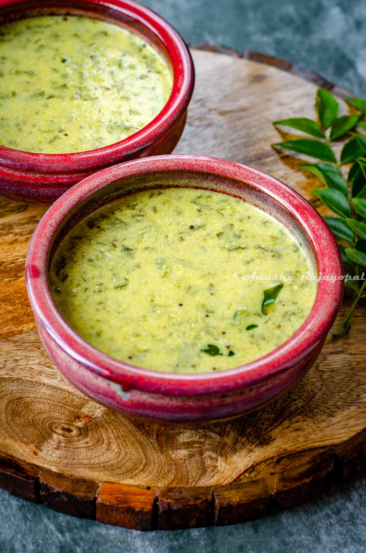 South Indian Mor Kulambu served in a reddish bowl placed over a wooden platter. Curry leaves at the background.