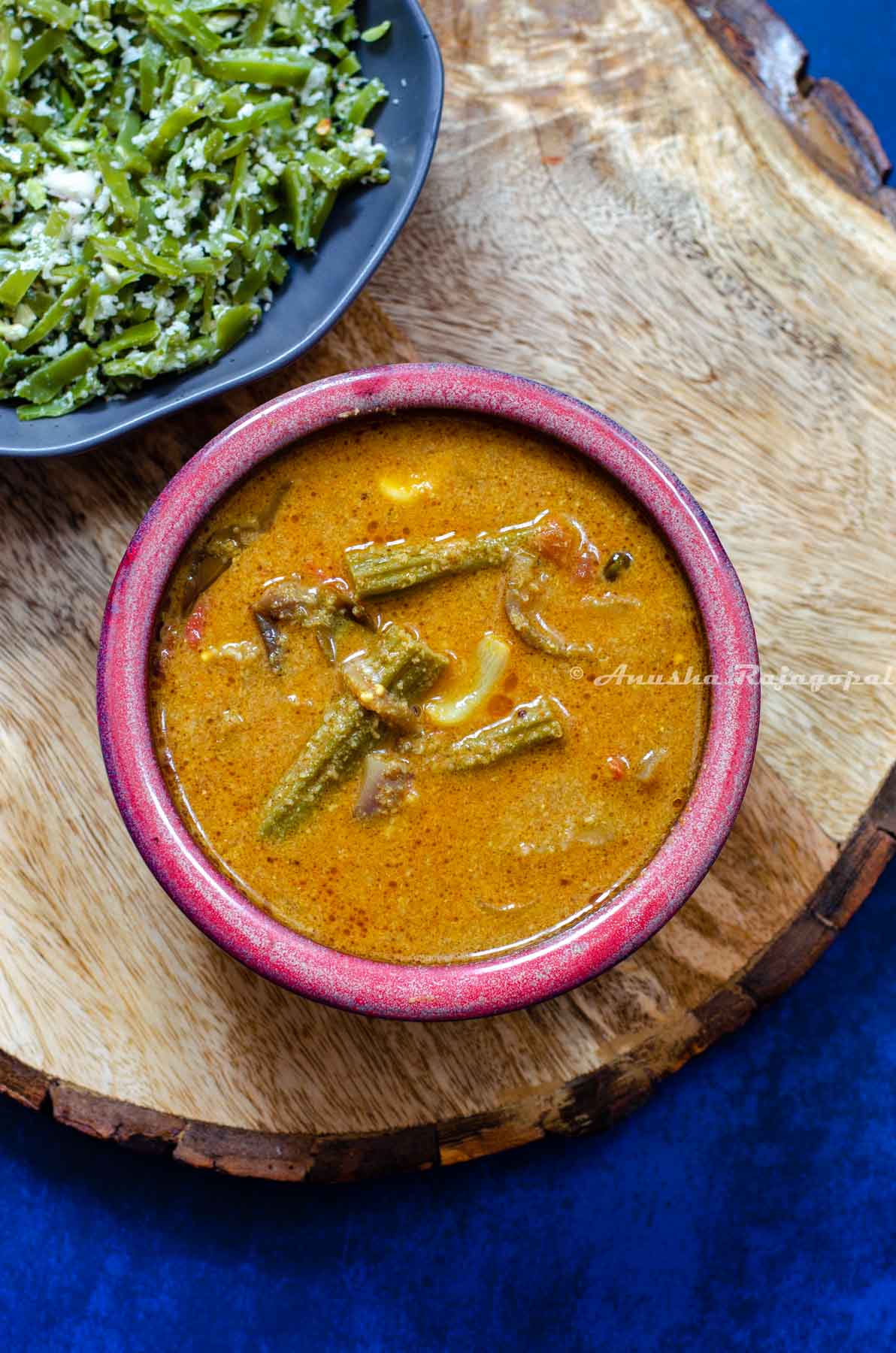 kara kulambu in a red bowl served with broad beans stir fry in a grey bowl. Both bowls on a wooden board