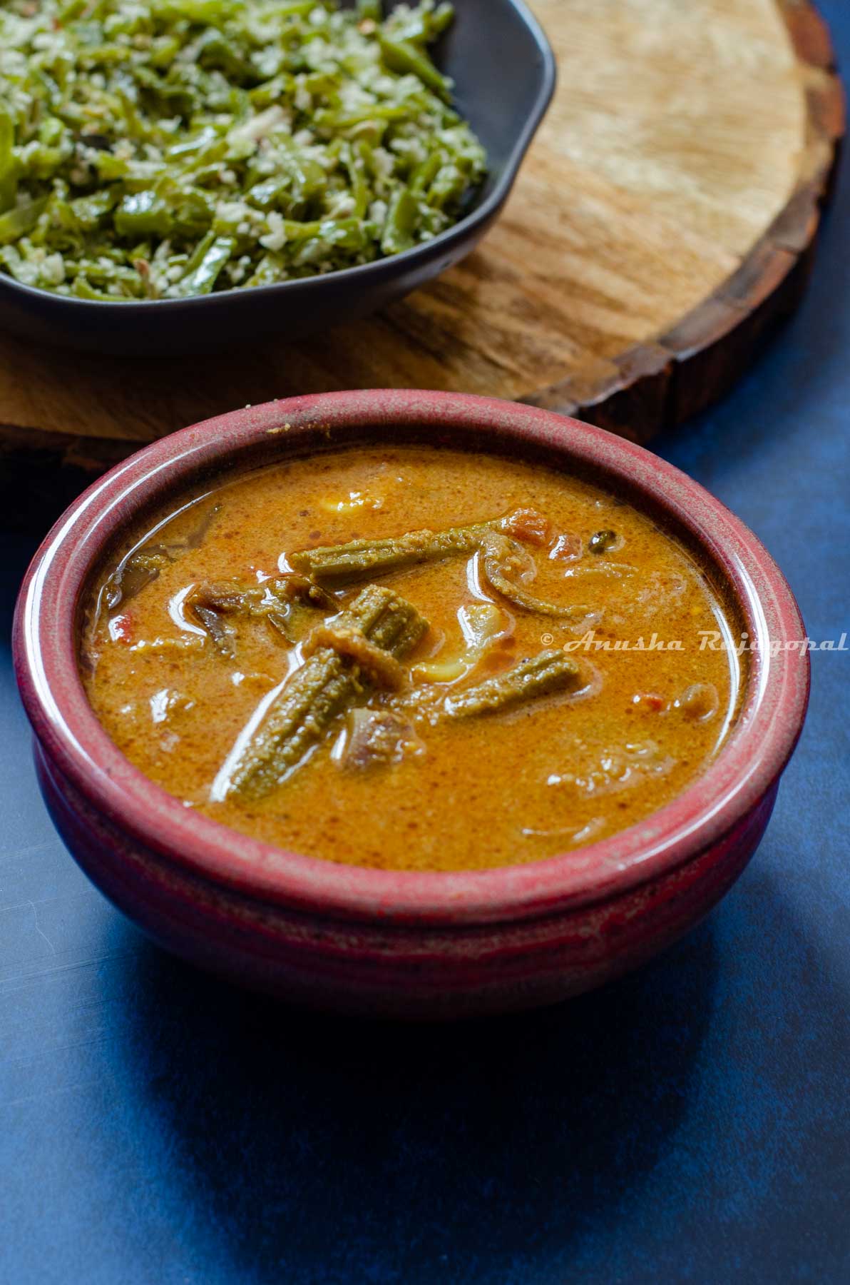 At the foregroud, a bowl of kara kulambu. Featured in the background is a bowl of broad beans stir fry placed over a wooden board.