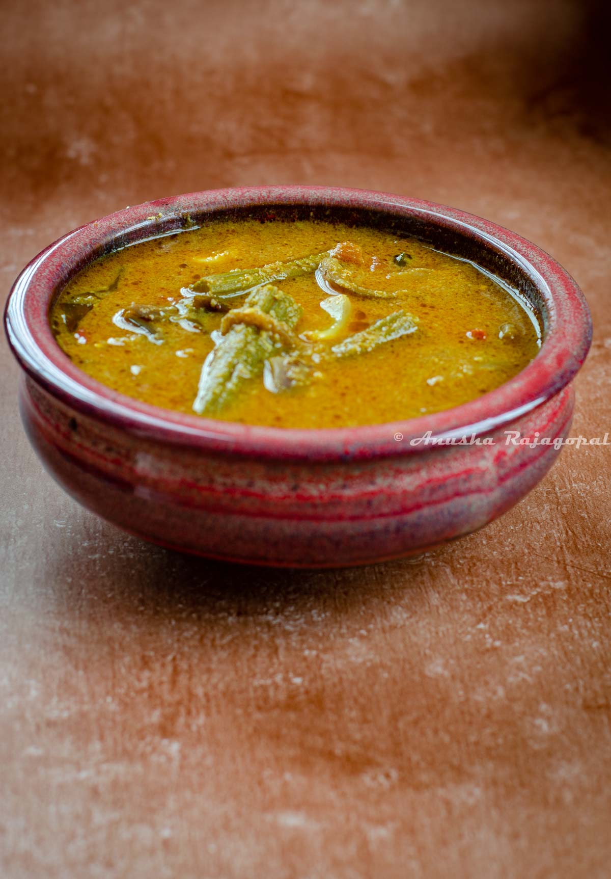 kara kulambu, a tangy gravy with eggplants and moringa served in a rustic red bowl placed over a brown backdrop