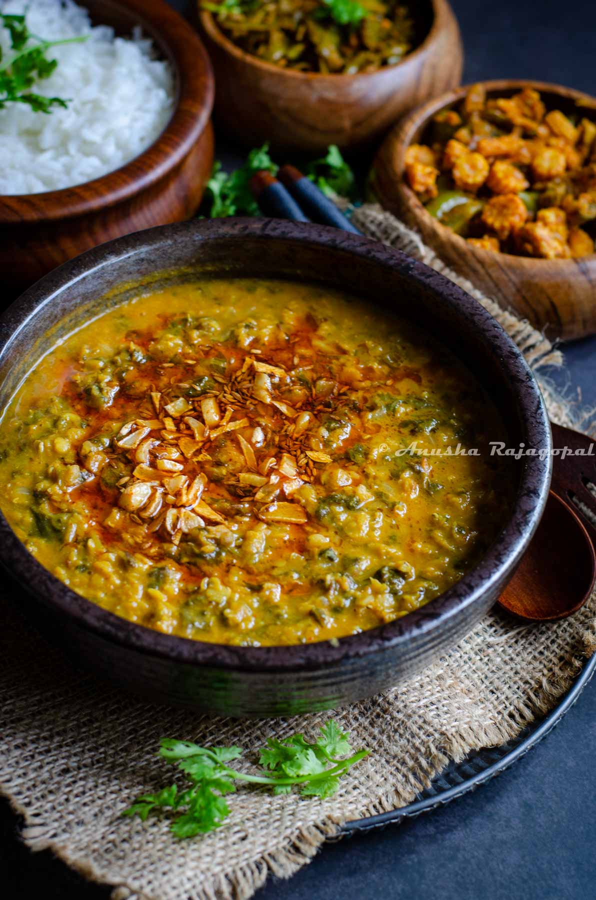 Kale dal served with a tempering of fried garlic and red paprika on top. The bowl of dal is placed over a burlap map. Rice and veggies served in wooden bowls by the side.