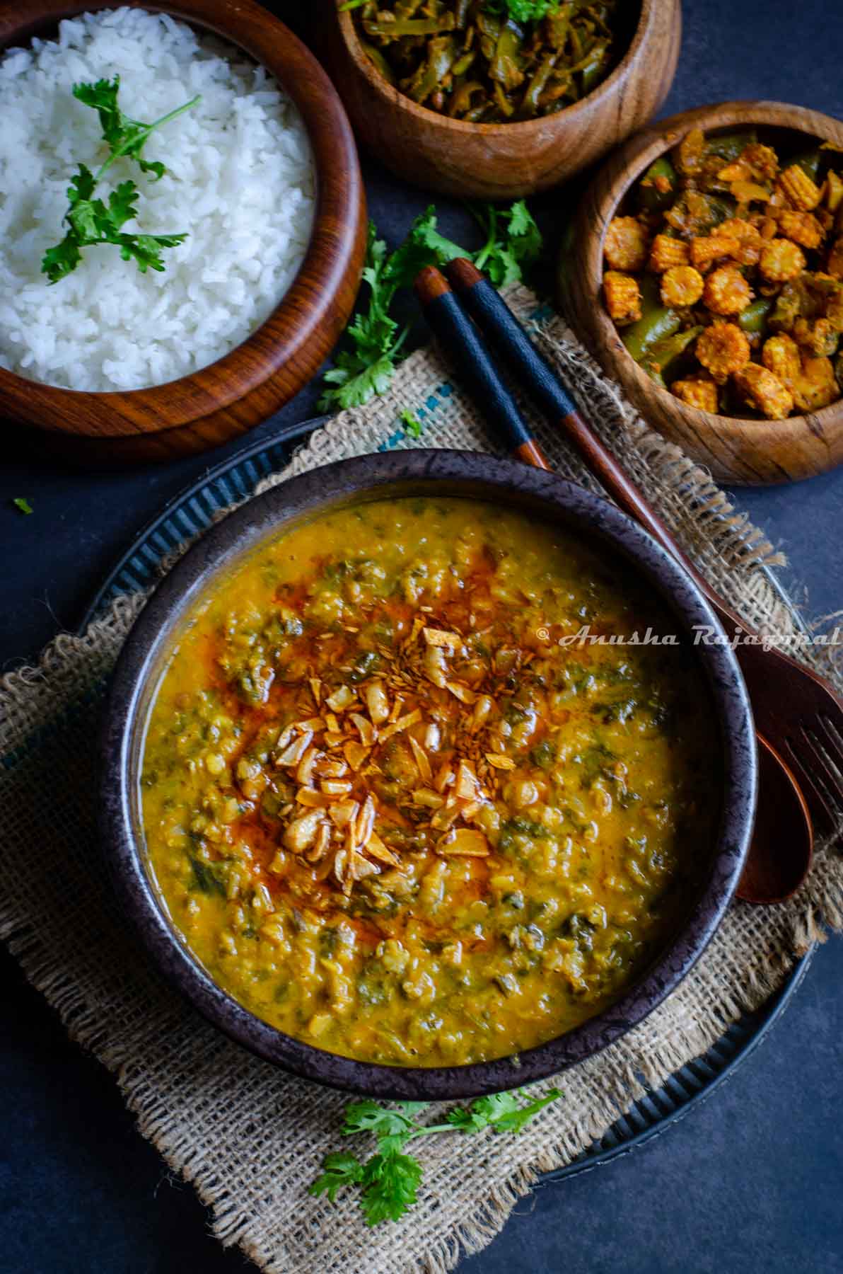 Kale dal served with a tempering of fried garlic and red paprika on top. The bowl of dal is placed over a burlap map. Rice and veggies served in wooden bowls by the side.