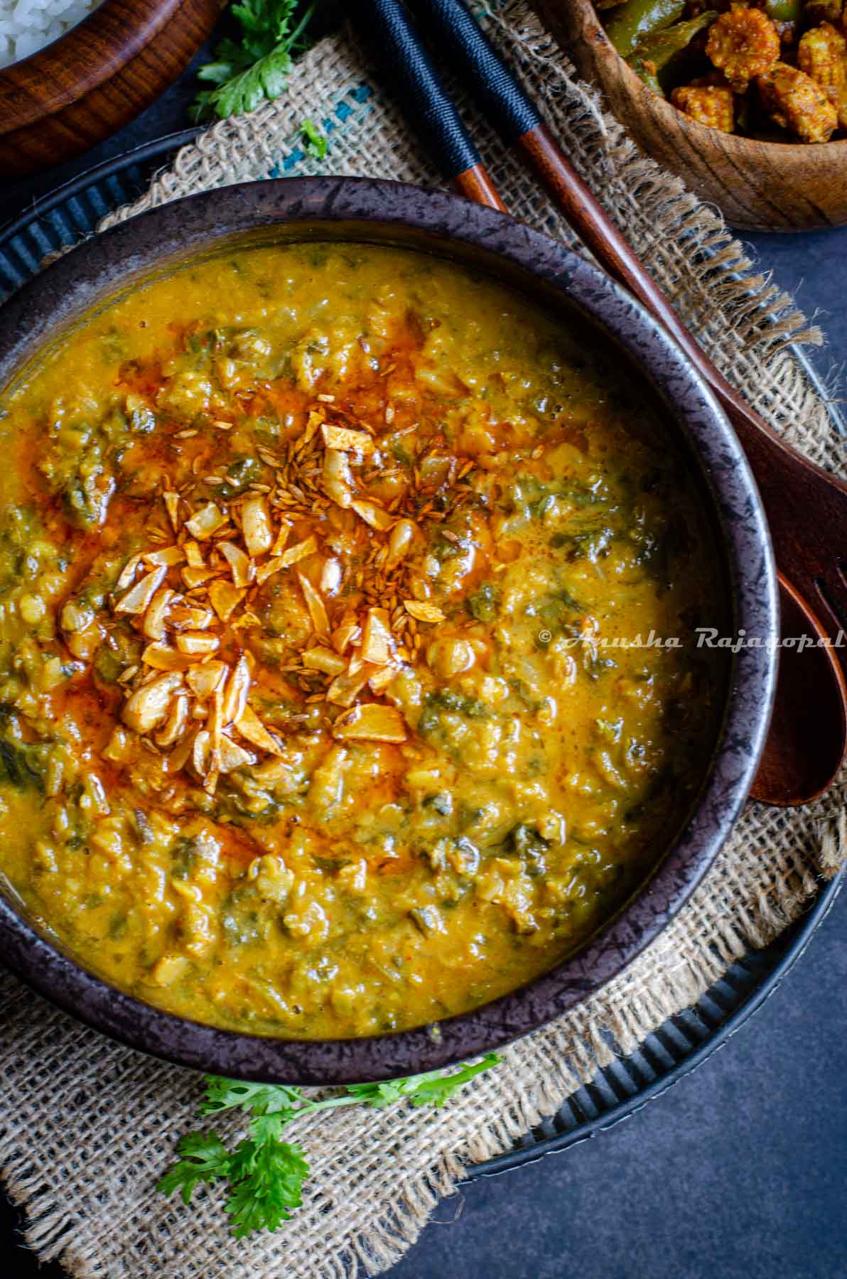 Kale dal served with a tempering of fried garlic and red paprika on top. The bowl of dal is placed over a burlap map. Rice and veggies served in wooden bowls by the side.