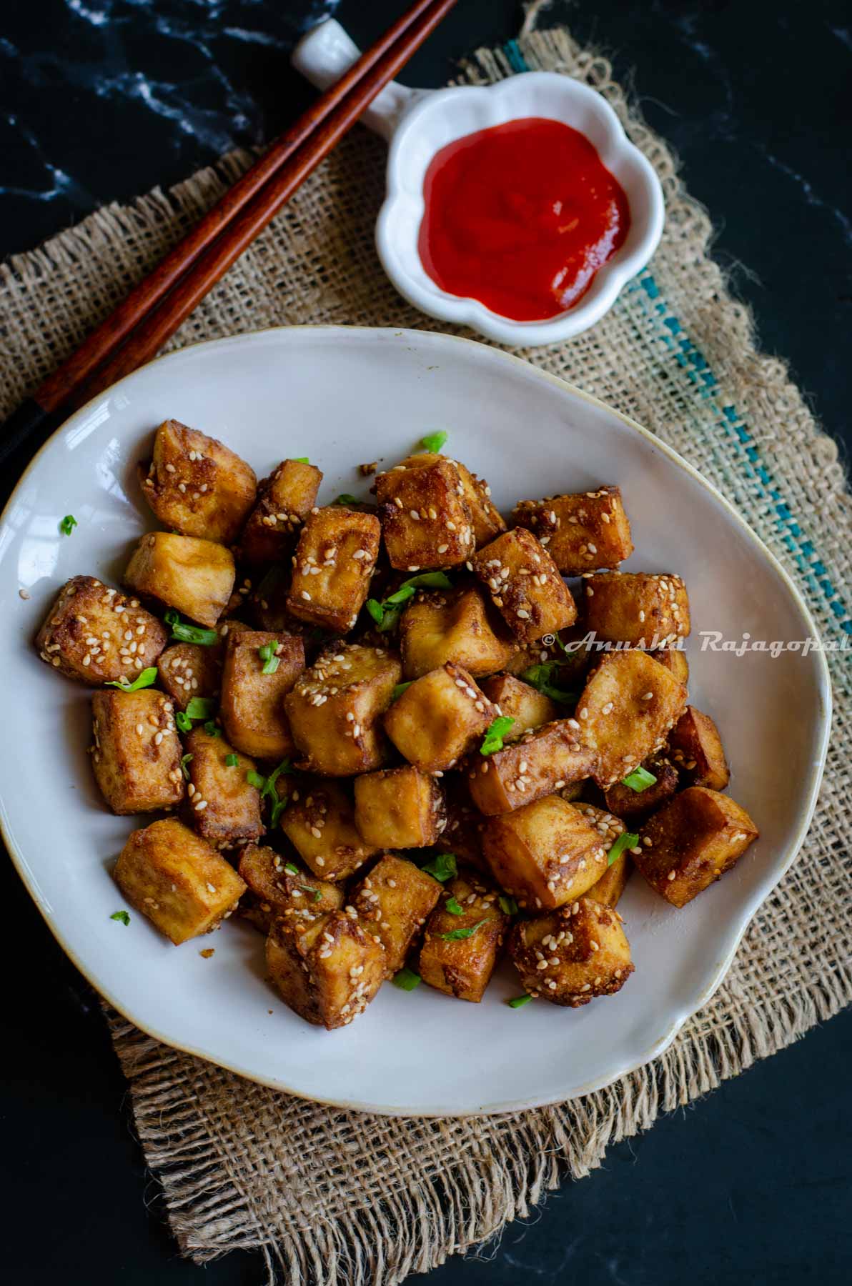 air fried tofu served in an asymmetric rimmed bowl with red chili sauce. chopsticks by the bowl