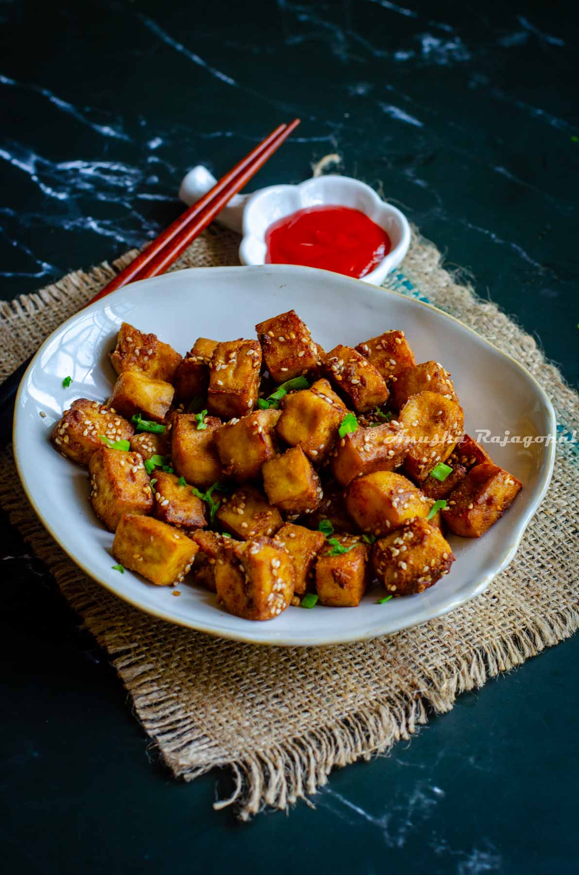 air fried tofu served in an asymmetric rimmed bowl with red chili sauce. chopsticks on the bowl.