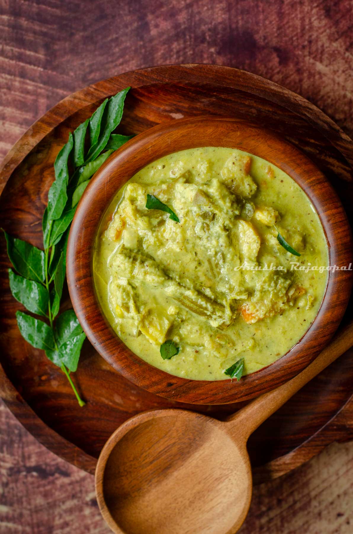 Kerala aviyal served in a wooden bowl placed over a wooden platter. A serving ladle and a sprig of curry leaves placed around the bowl.