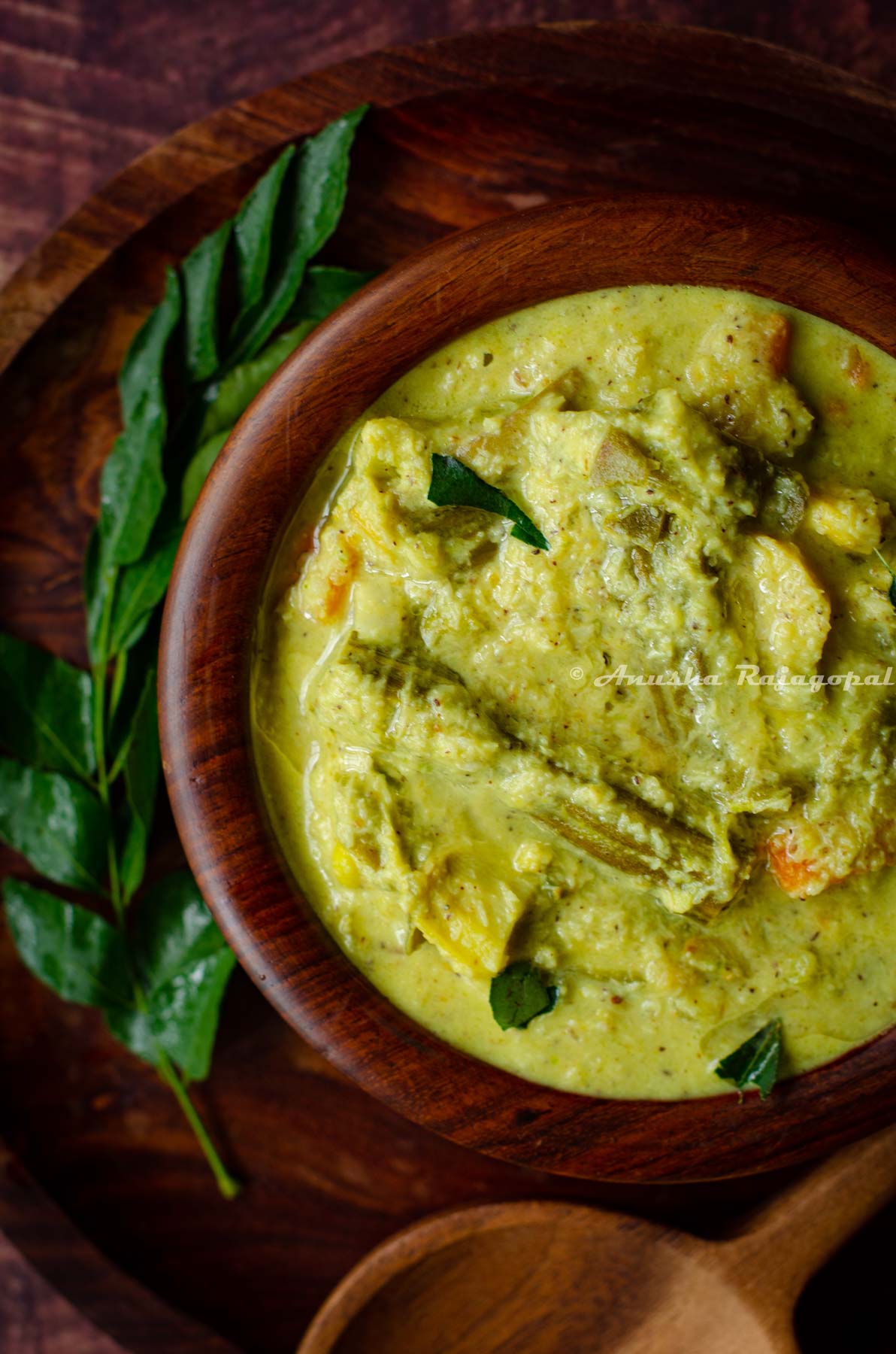 Kerala aviyal served in a wooden bowl placed over a wooden platter. A serving ladle and a sprig of curry leaves placed around the bowl.