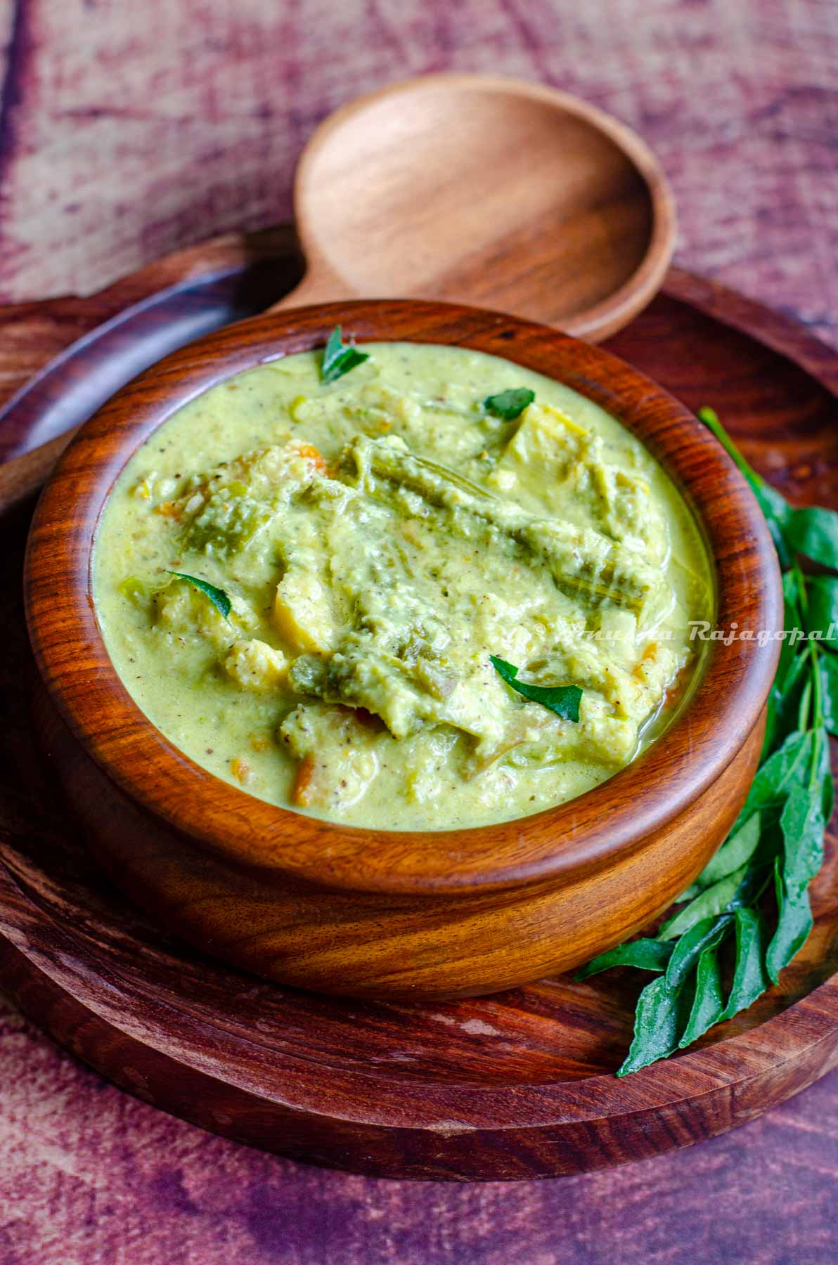 Kerala aviyal served in a wooden bowl placed over a wooden platter. A serving ladle and a sprig of curry leaves placed around the bowl.