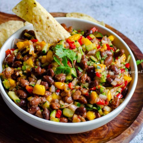 black bean and mango salad served in a white shallow bowl with a tortilla chip inserted on the top. The bowl has been placed over a wooden platter with a handle. Tortilla chips lay scattered behind the bowl.