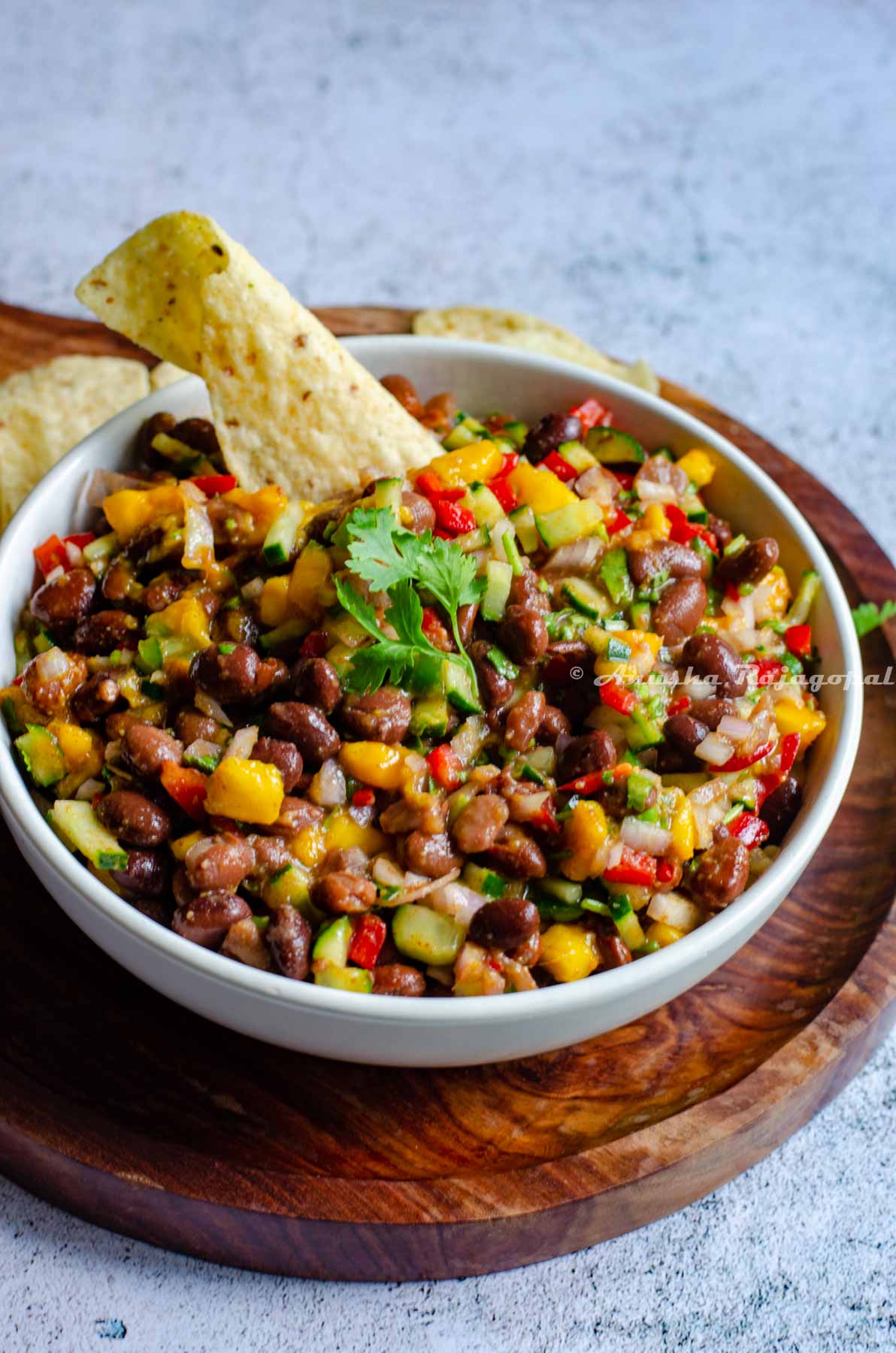 black bean and mango salad served in a white shallow bowl with a tortilla chip  inserted on the top. The bowl has been placed over a wooden platter with a handle. Tortilla chips lay scattered behind the bowl.