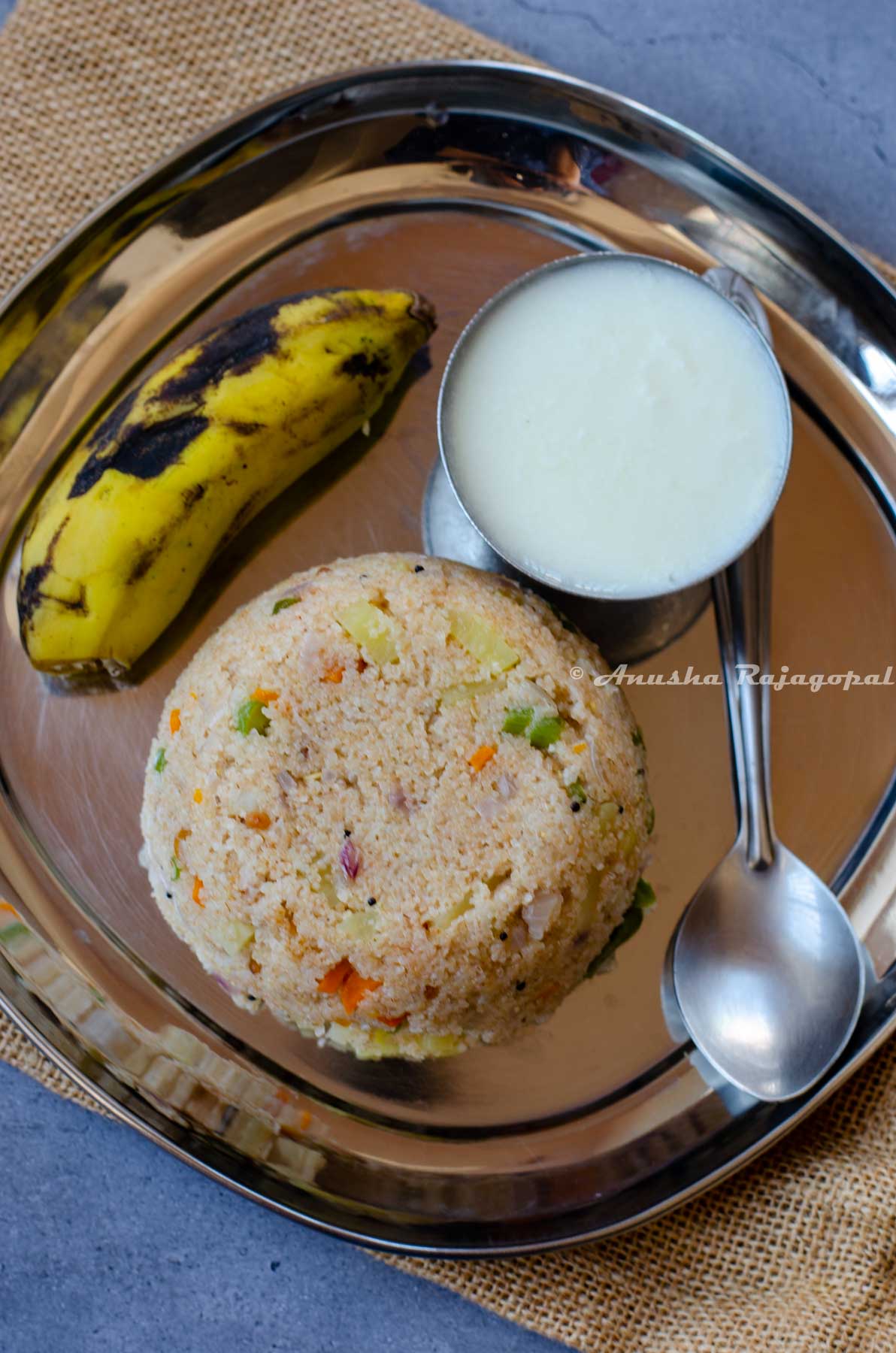 broken wheat upma served on a steel plate with a banana, yogurt and a spoon by the side.