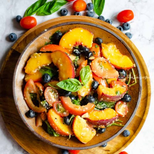 vegan peach salad with summer fruit served in Khaki brown shallow bowl placed over a wooden plate. Fruits scattered around the plate.