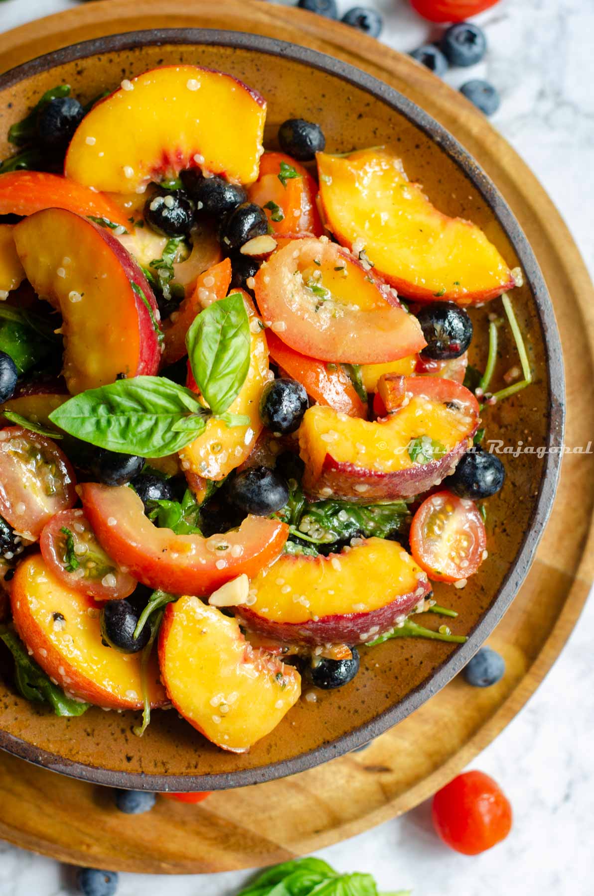 vegan peach salad with summer fruit served in Khaki brown shallow bowl placed over a wooden plate. Fruits scattered around the plate.